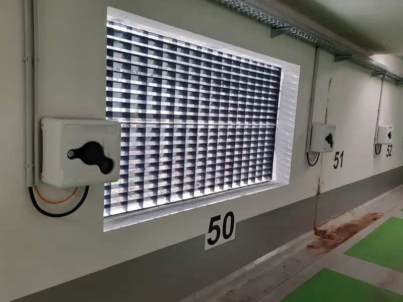 Parking souterrain : un boîtier de recharge blanc fixé au mur de la place 50, alimenté par un conduit qui descend le long du mur, à côté d’une grille d’aération