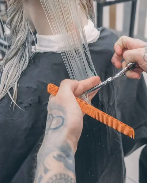 Mains tatouées qui coupent une mèche blond polaire avec un peigne orange