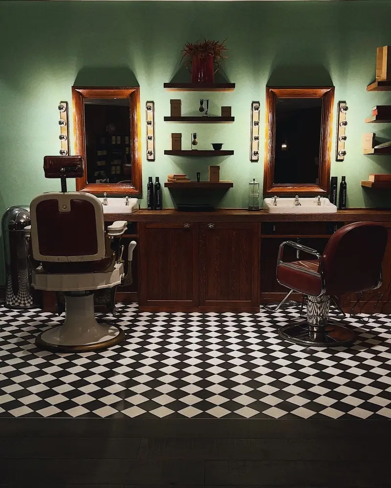 Fauteuils de barbier en cuir, miroirs encadrés de bois et carrelage noir et blanc sur un mur vert
