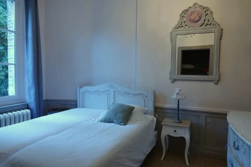 Chambre aux murs gris-bleu, deux lits accolés faits de draps blancs, table de chevet et miroir ancien