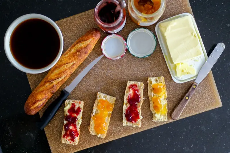 Tartines beurrées à la confiture d’abricot et de fraise, baguette, bol de café, pots de confiture