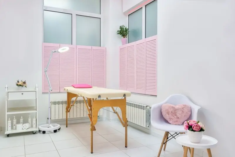 Cabine claire avec table de soin en bois, volets roses, lampe loupe et fauteuil blanc