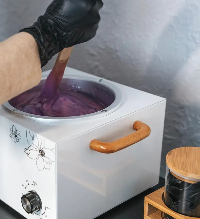 Main gantée de noir qui remue la cire tiède violette dans un chauffe-cire blanc