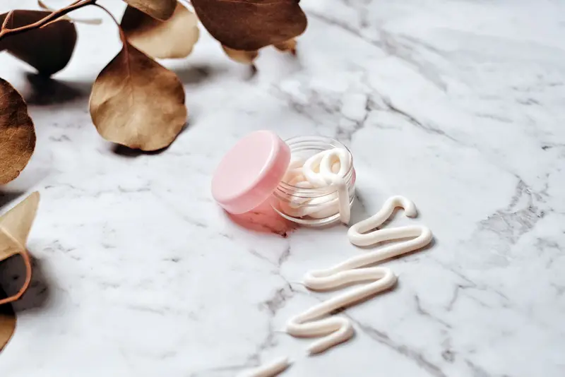 Pot de crème rose ouvert et traits de crème sur un plateau de marbre blanc, entre des feuilles séchées