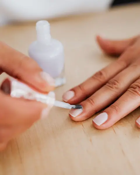 Pinceau qui pose un vernis lilas pâle sur une main à plat, sur une table en bois clair