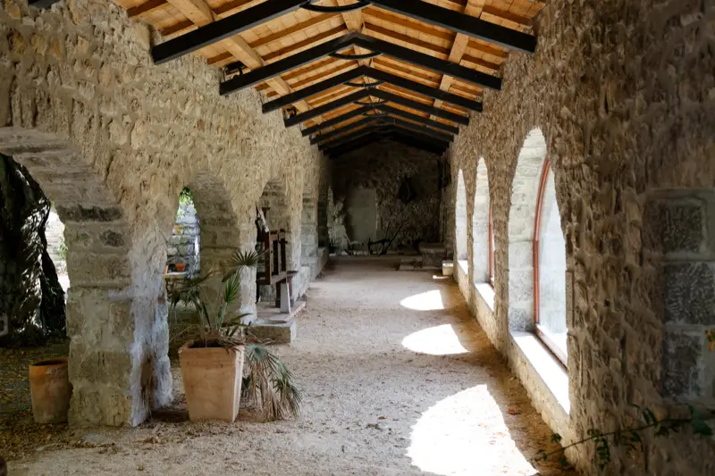 Longue galerie aux murs de pierre, des arcades d’un côté et de hautes fenêtres cintrées de l’autre, sous une charpente de bois clair, sol de gravier et palmiers en pot