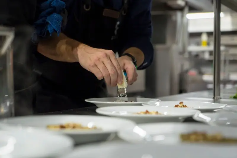 Mains d’un cuisinier qui râpe du fromage sur une rangée d’assiettes, sur le passe de la cuisine
