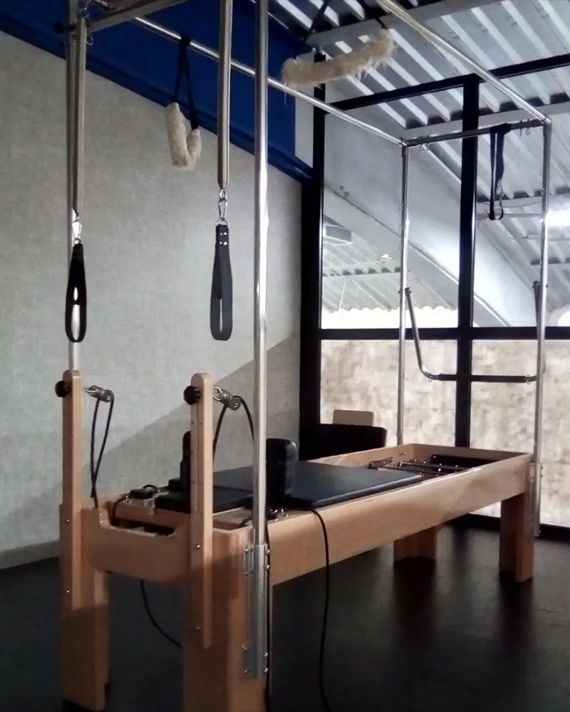 Reformer en bois clair sous un cadre en acier d’où pendent des sangles, devant une verrière d’atelier