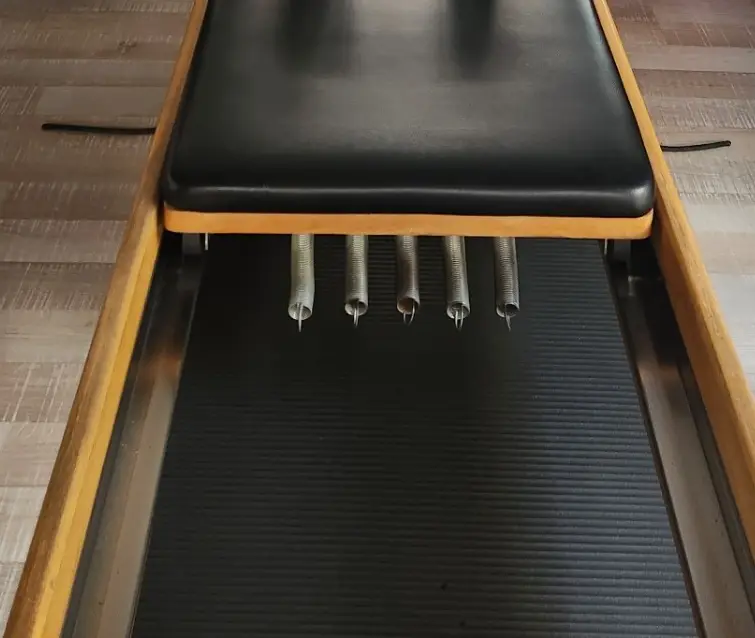 Chariot de reformer vu de face, rembourrage noir, cinq ressorts accrochés dessous, rails en bois de chaque côté